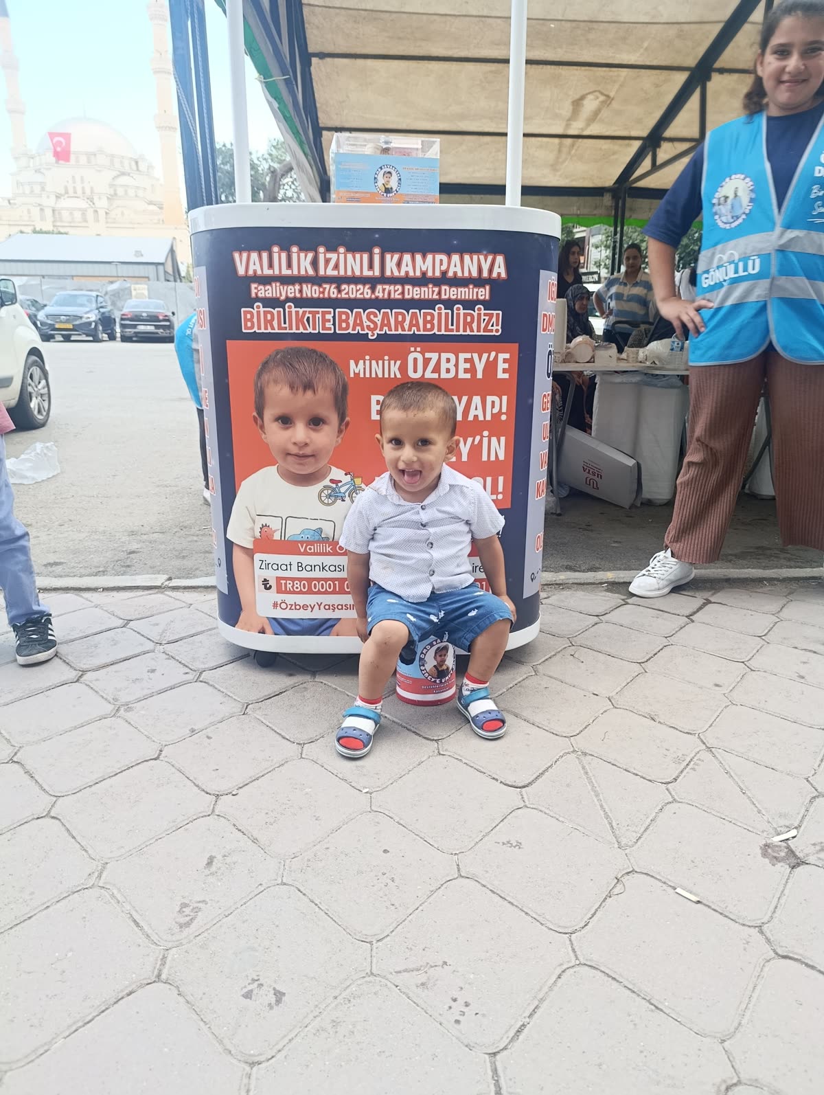 Özbey, valilik izinli kampanya standının önünde kumbaranın üzerinde oturmuş gülümsüyor