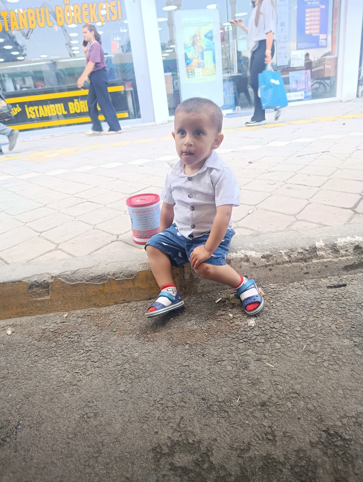 Özbey kaldırımda oturuyor, yanında kampanya kumbarası